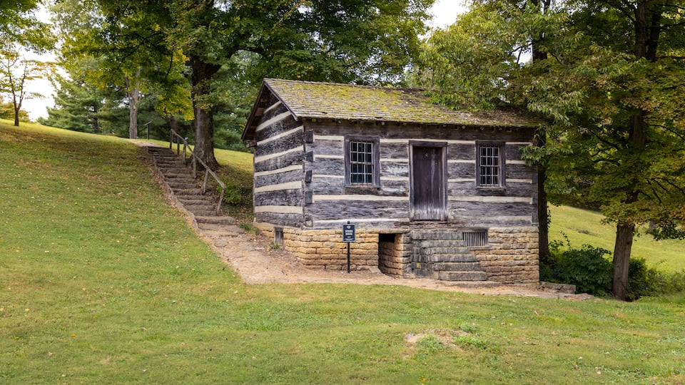 A log cabin with a stone foundation and a wooden door, surrounded by trees and a grassy hill.