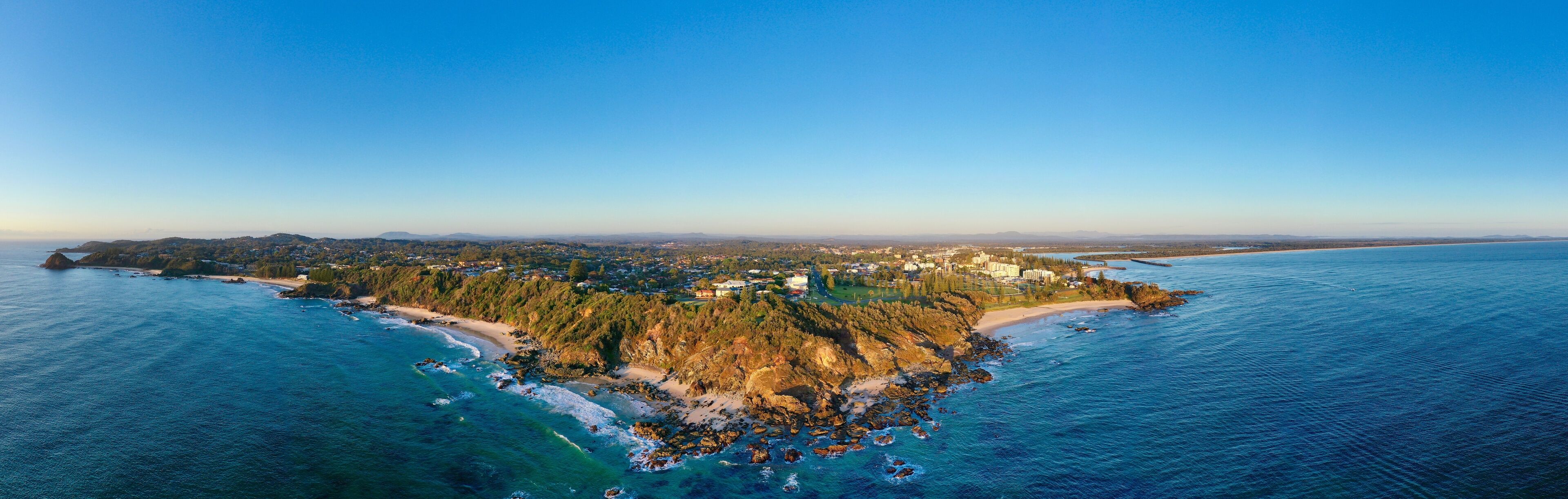An aerial view of a coastal town with a beach, forested area, and a clear blue sky.
