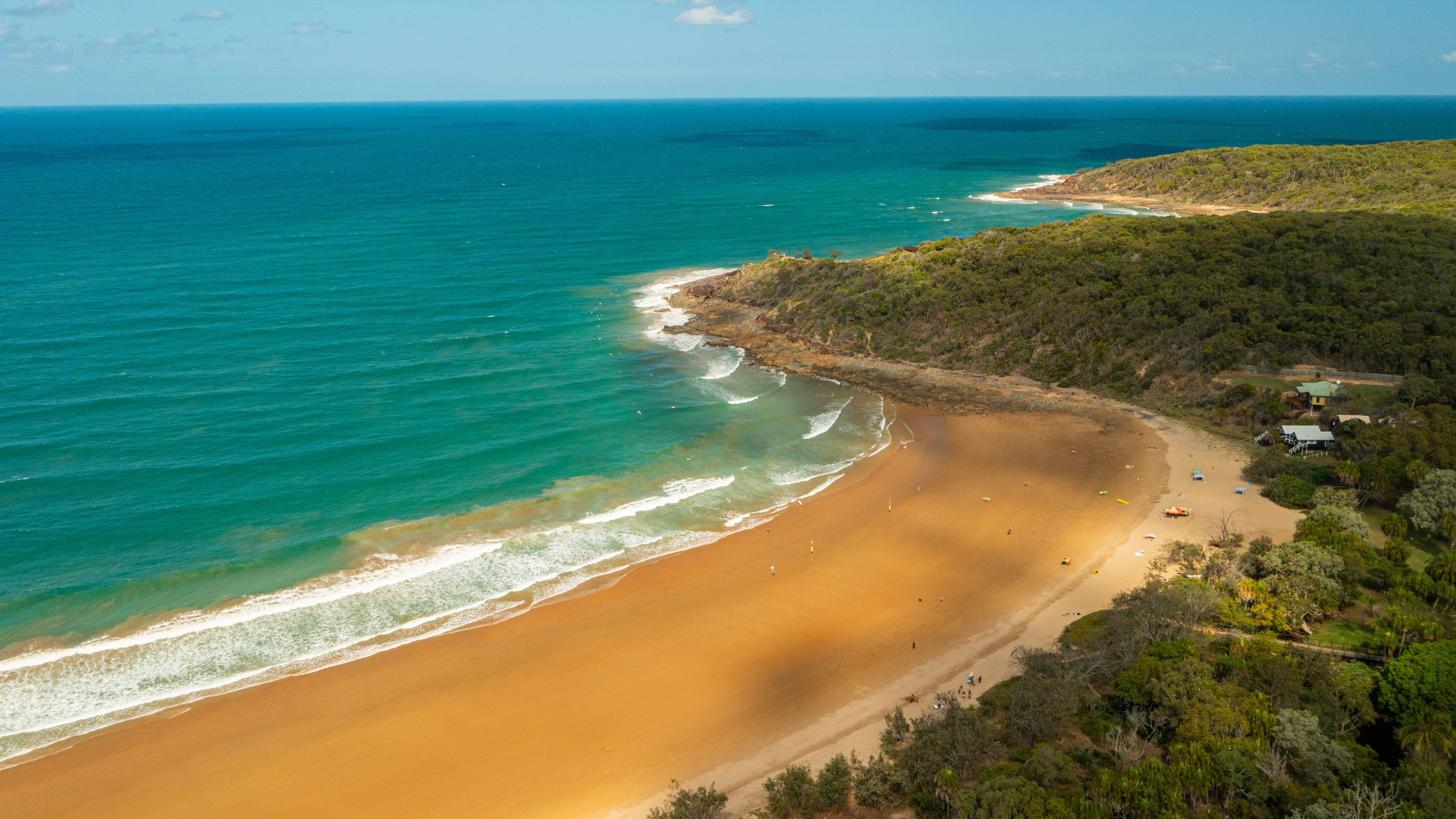 A coastal landscape with a sandy beach, turquoise waters, and a forested area with buildings.