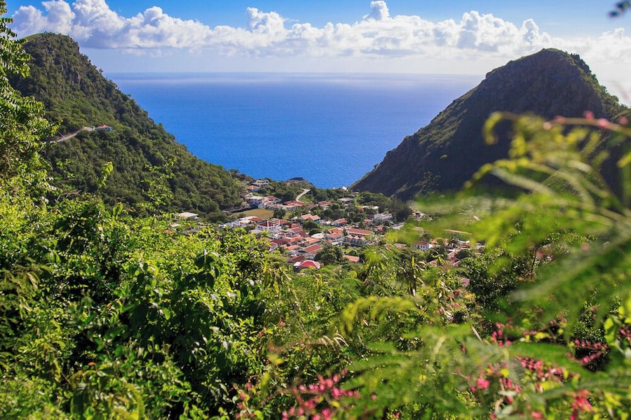 A coastal town nestled between lush green hills, with a clear view of the ocean and a winding road leading to the settlement.