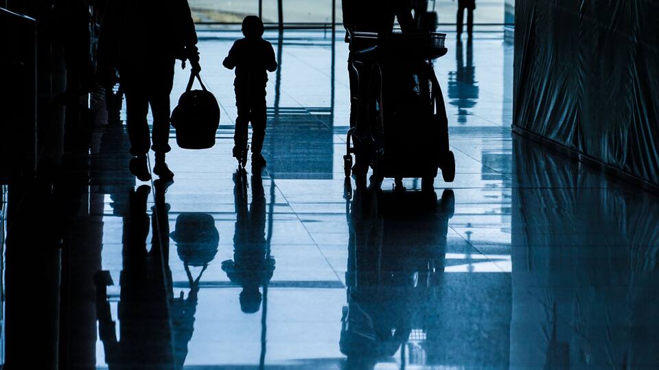 Silhouettes of people walking in a reflective corridor with a shiny floor and a large, dark structure on the right.