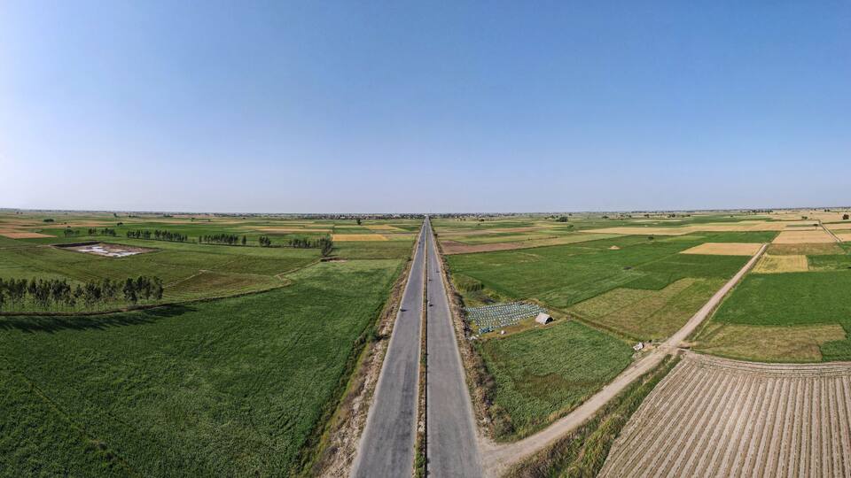 A straight road through a rural landscape with green fields and a clear sky.