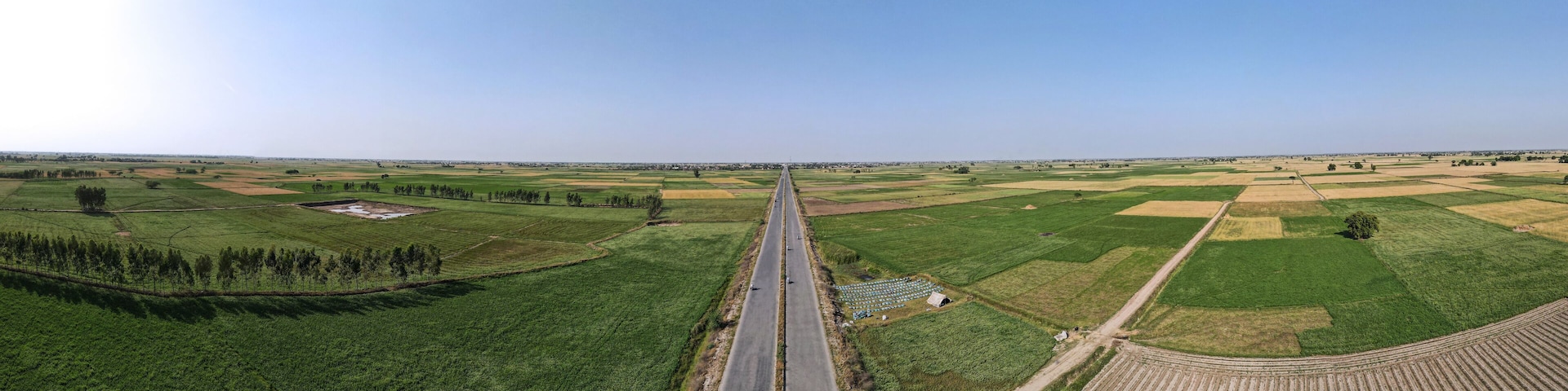 A straight road through a rural landscape with green fields and a clear sky.