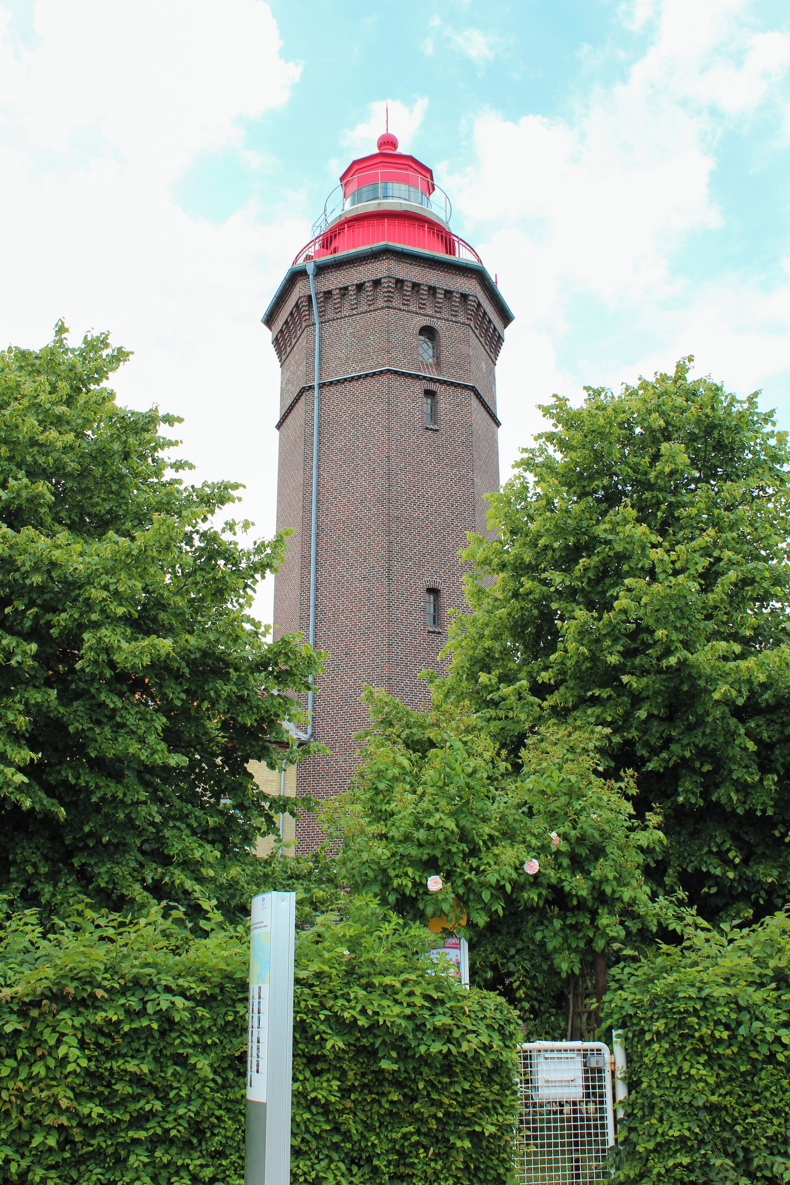A brick lighthouse with a red top, surrounded by lush green trees.