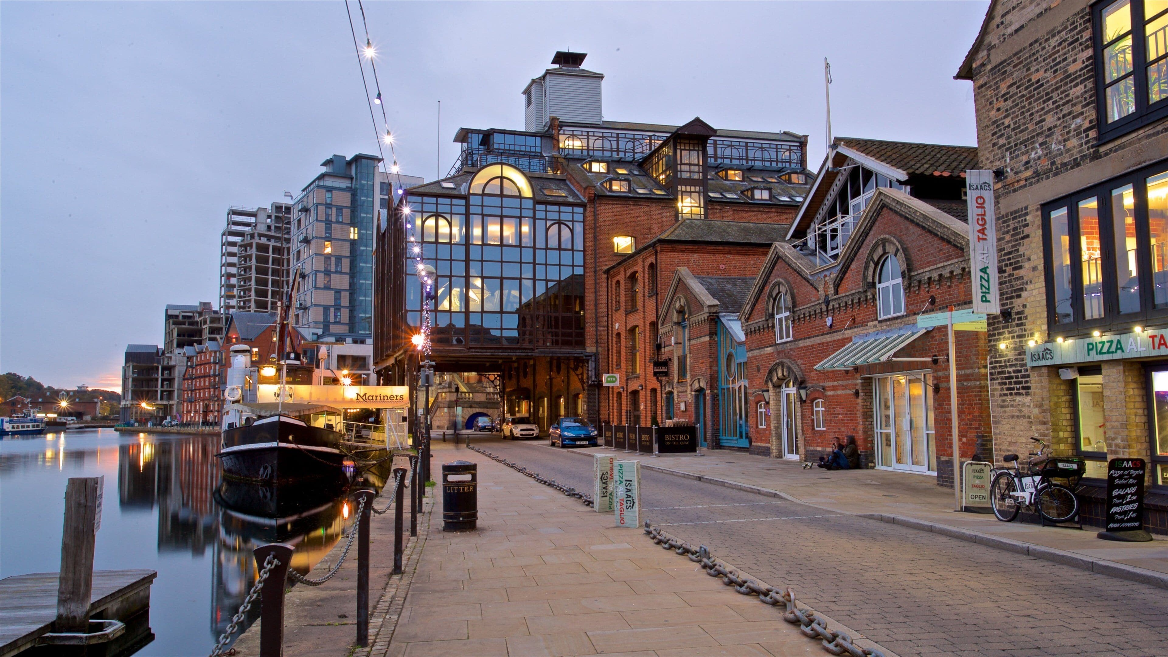 A waterfront with a marina, a modern building with large windows, and a brick building with a sign for 'Pizza Al Taglio'.