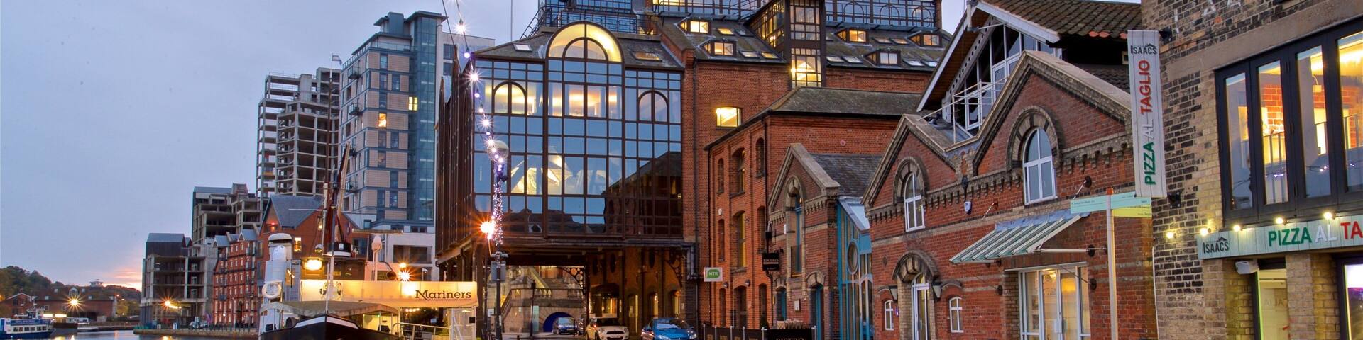 A waterfront with a marina, a modern building with large windows, and a brick building with a sign for 'Pizza Al Taglio'.