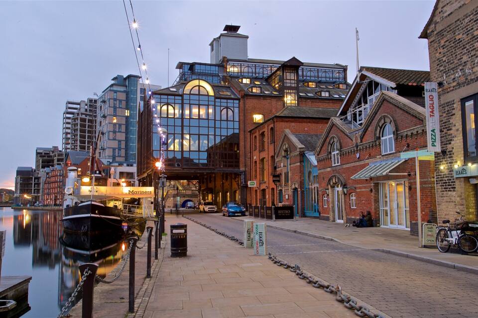 A waterfront with a marina, a modern building with large windows, and a brick building with a sign for 'Pizza Al Taglio'.