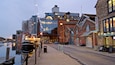 A waterfront with a marina, a modern building with large windows, and a brick building with a sign for 'Pizza Al Taglio'.