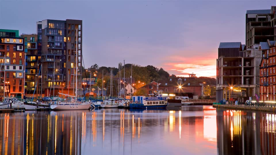 A marina with boats docked, surrounded by modern buildings and a sunset in the background.