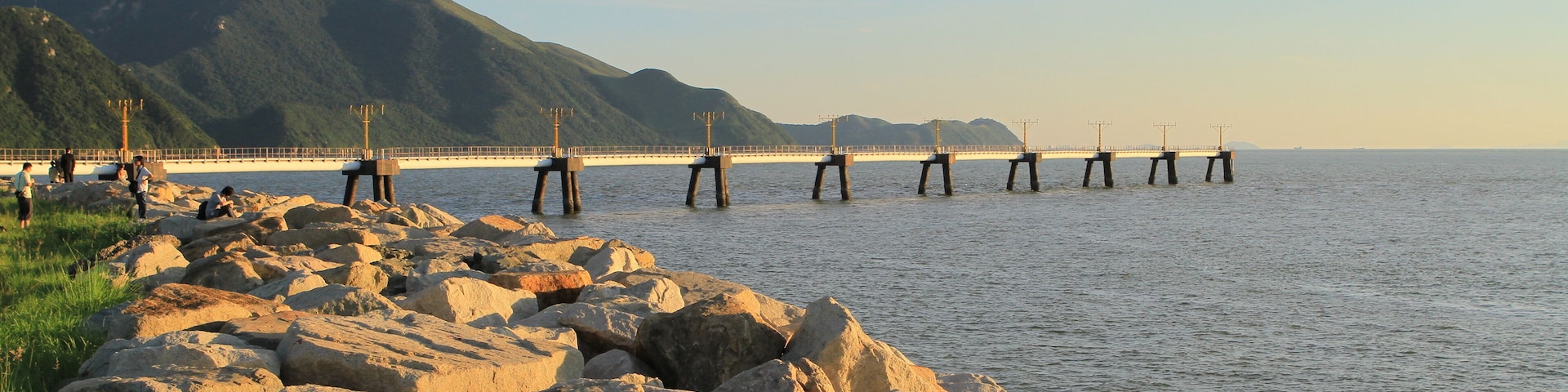 Una costa rocosa con un puente que se adentra en el agua, rodeada de montañas.