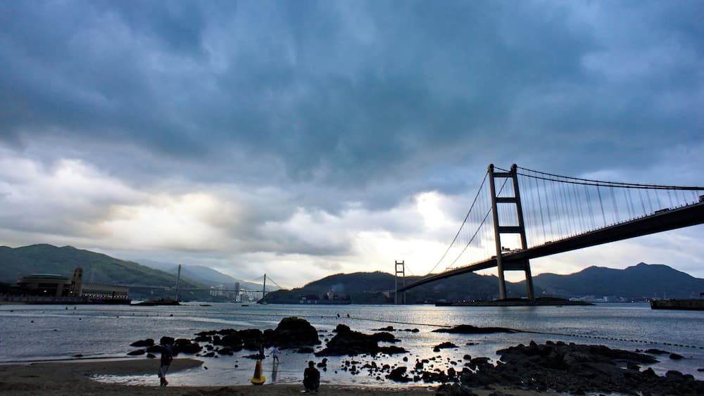 A suspension bridge over a river with rocky shores and mountains in the background.