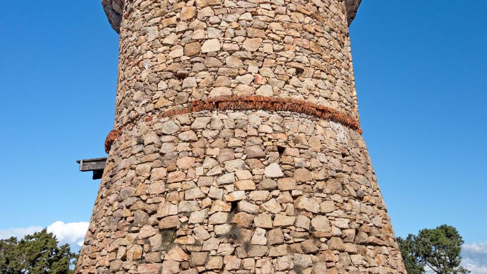 A stone tower with a circular top, surrounded by a clear blue sky and a distant view of the sea.