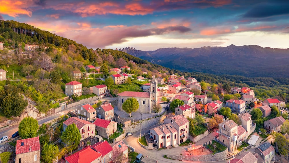 Un village aux bâtiments aux toits rouges, niché dans une vallée, entouré de montagnes et d’une végétation luxuriante.