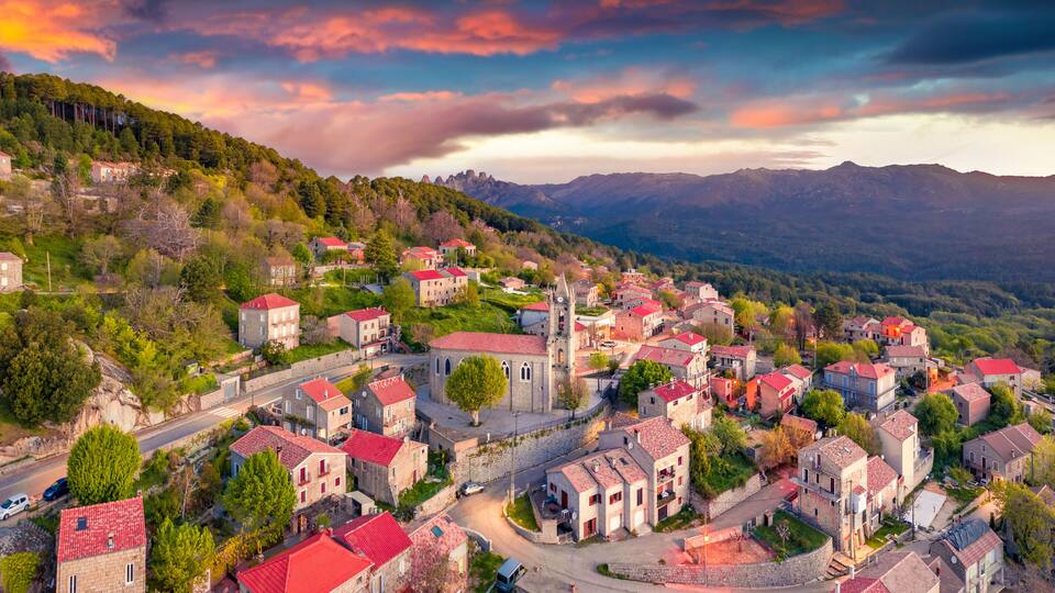 A village with red-roofed buildings nestled in a valley, surrounded by mountains and lush greenery.
