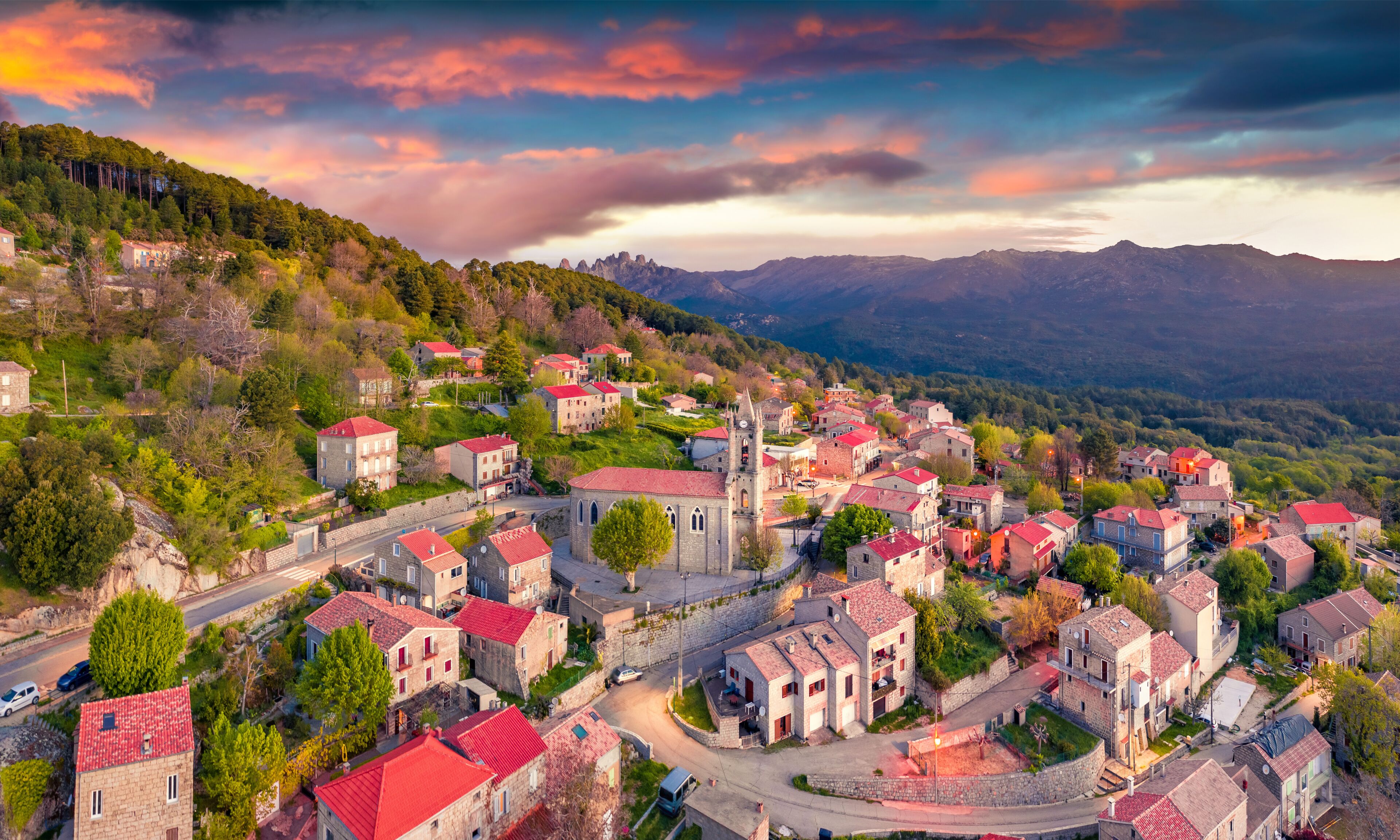 A village with red-roofed buildings nestled in a valley, surrounded by mountains and lush greenery.