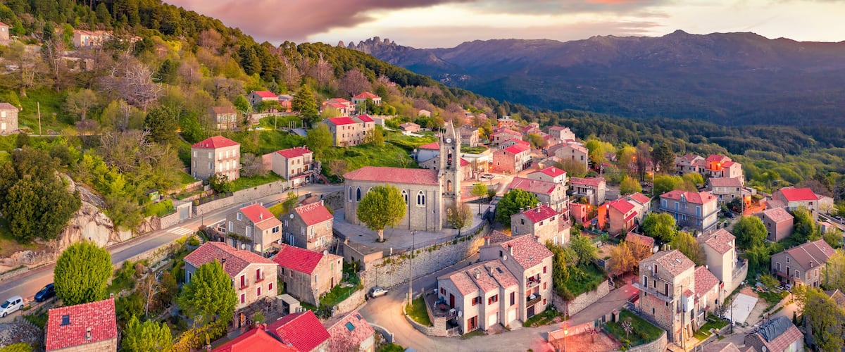 A village with red-roofed buildings nestled in a valley, surrounded by mountains and lush greenery.