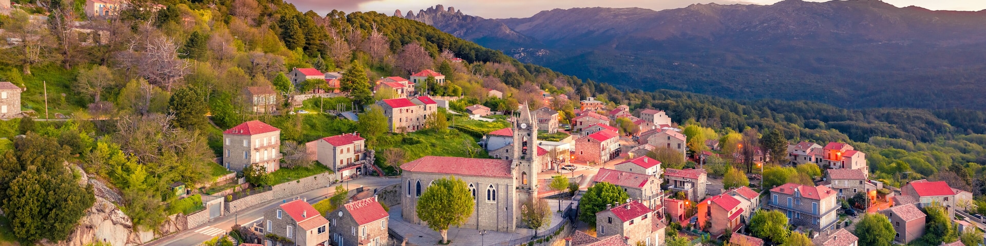 A village with red-roofed buildings nestled in a valley, surrounded by mountains and lush greenery.