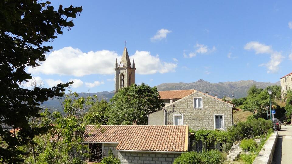 A stone church with a bell tower, surrounded by houses with tiled roofs and lush greenery.