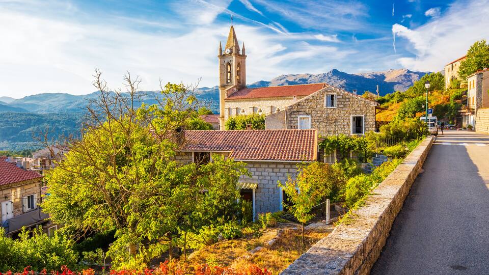 A stone church with a bell tower, surrounded by greenery and a mountain range in the background.