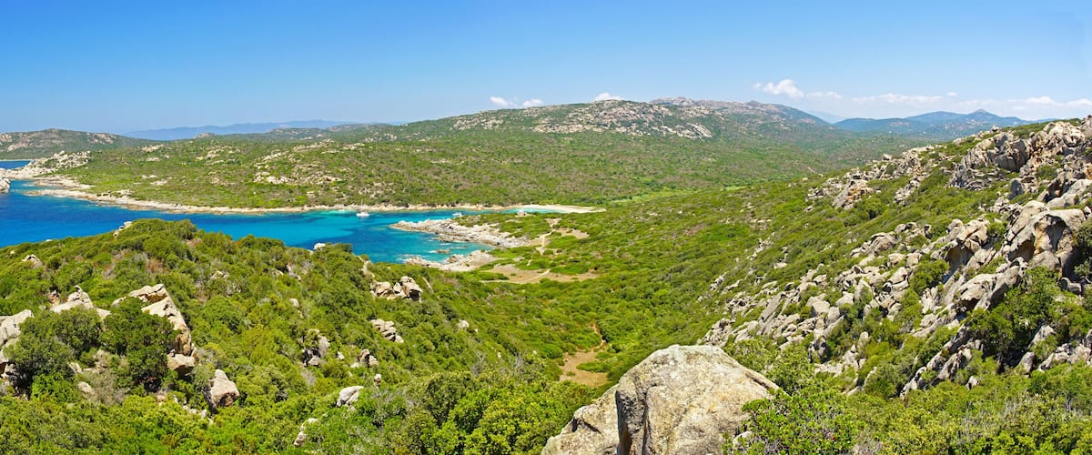 A coastal landscape with rocky terrain, dense greenery, and a clear blue sea.