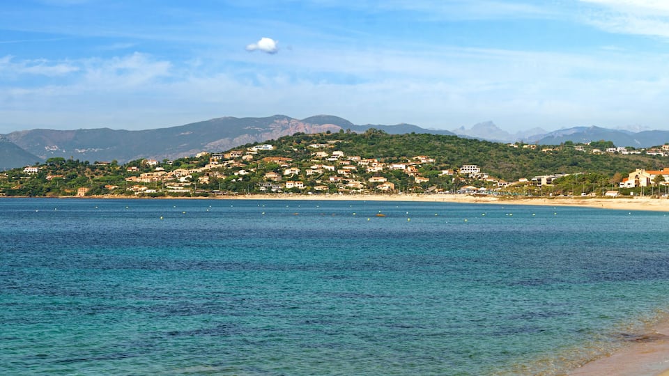 A coastal town with a beach, clear blue waters, and a mountain range in the background.