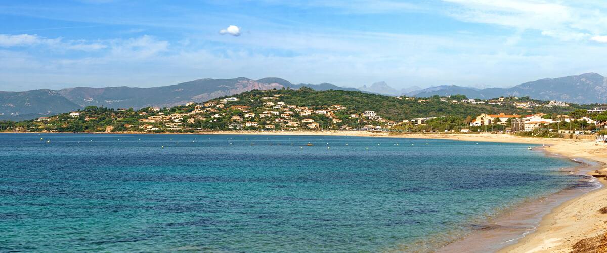 A coastal town with a beach, clear blue waters, and a mountain range in the background.