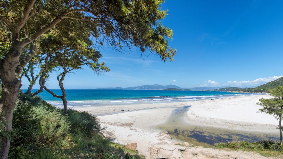 A coastal beach with clear blue waters, sandy shores, and green hills in the background.