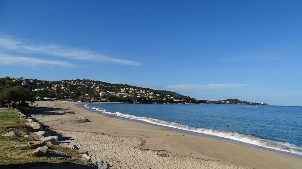 A beach with a rocky shoreline, clear blue water, and a hillside with houses in the background.