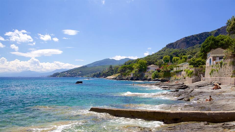 A coastal scene with clear blue waters, rocky shore, and buildings on the hillside.