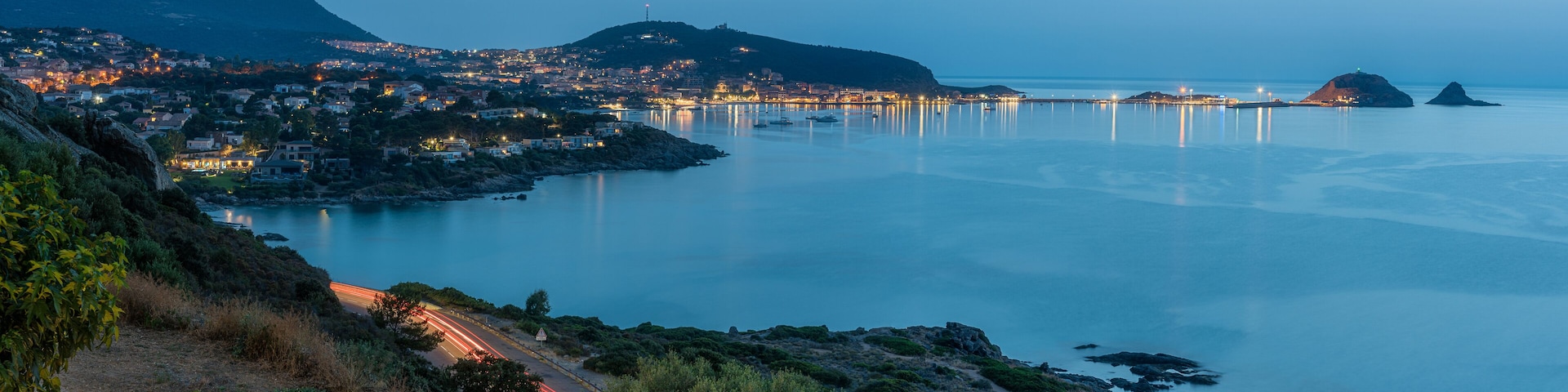 Uma vila costeira ao entardecer, com edifícios iluminados, mar calmo e uma colina imponente ao fundo.