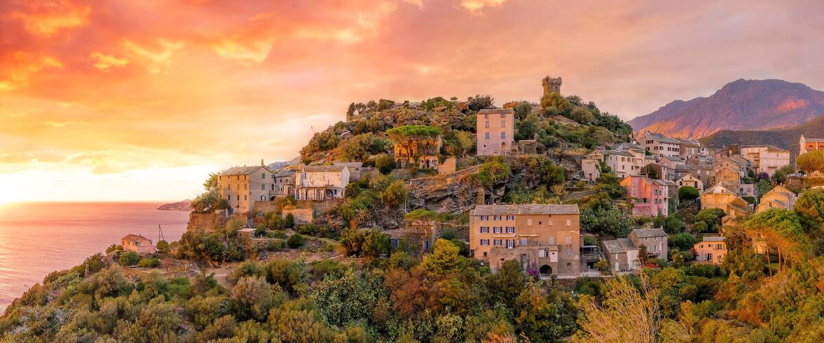 A coastal village with historic buildings, a prominent tower, and a sunset over the sea.