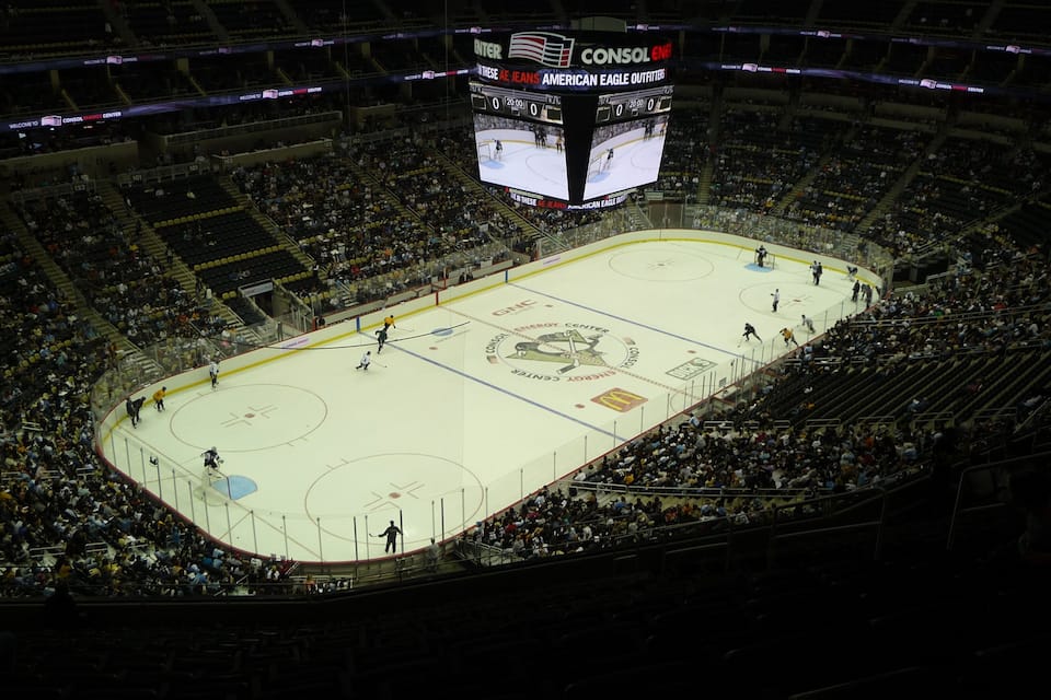 Une patinoire intérieure où se pratique le hockey sur glace, avec un tableau d’affichage, des gradins pour le public et des joueurs sur la glace.