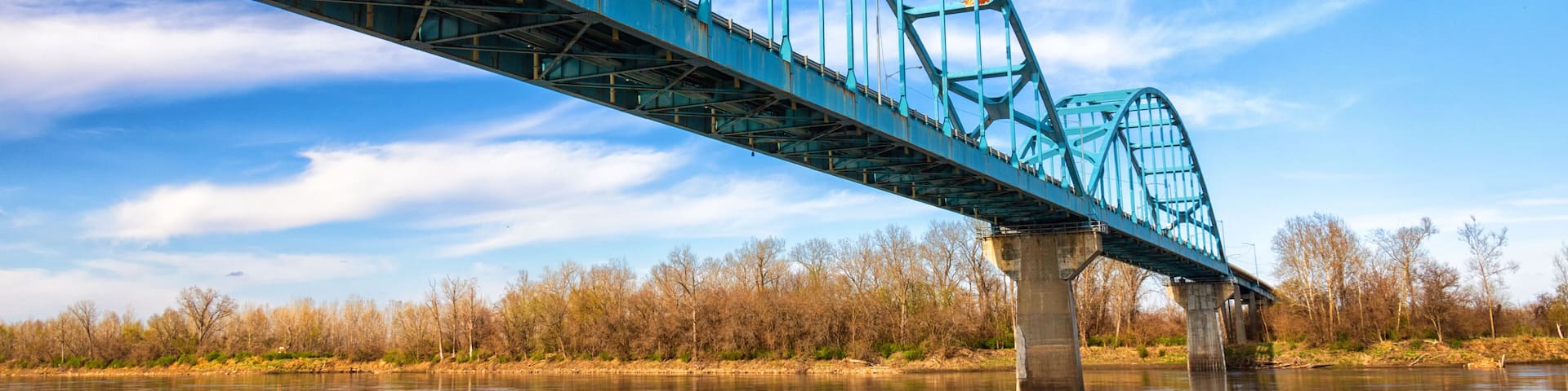 A blue steel bridge over a calm river with trees in the background.