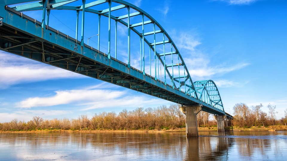 A blue steel bridge over a calm river with trees in the background.
