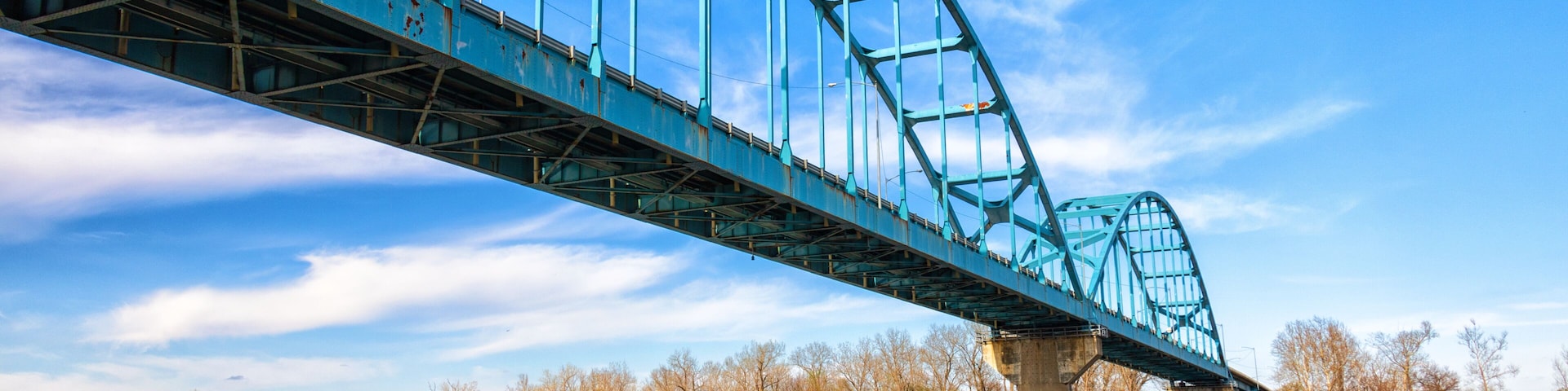 Un puente de acero azul sobre un río tranquilo, con árboles al fondo.