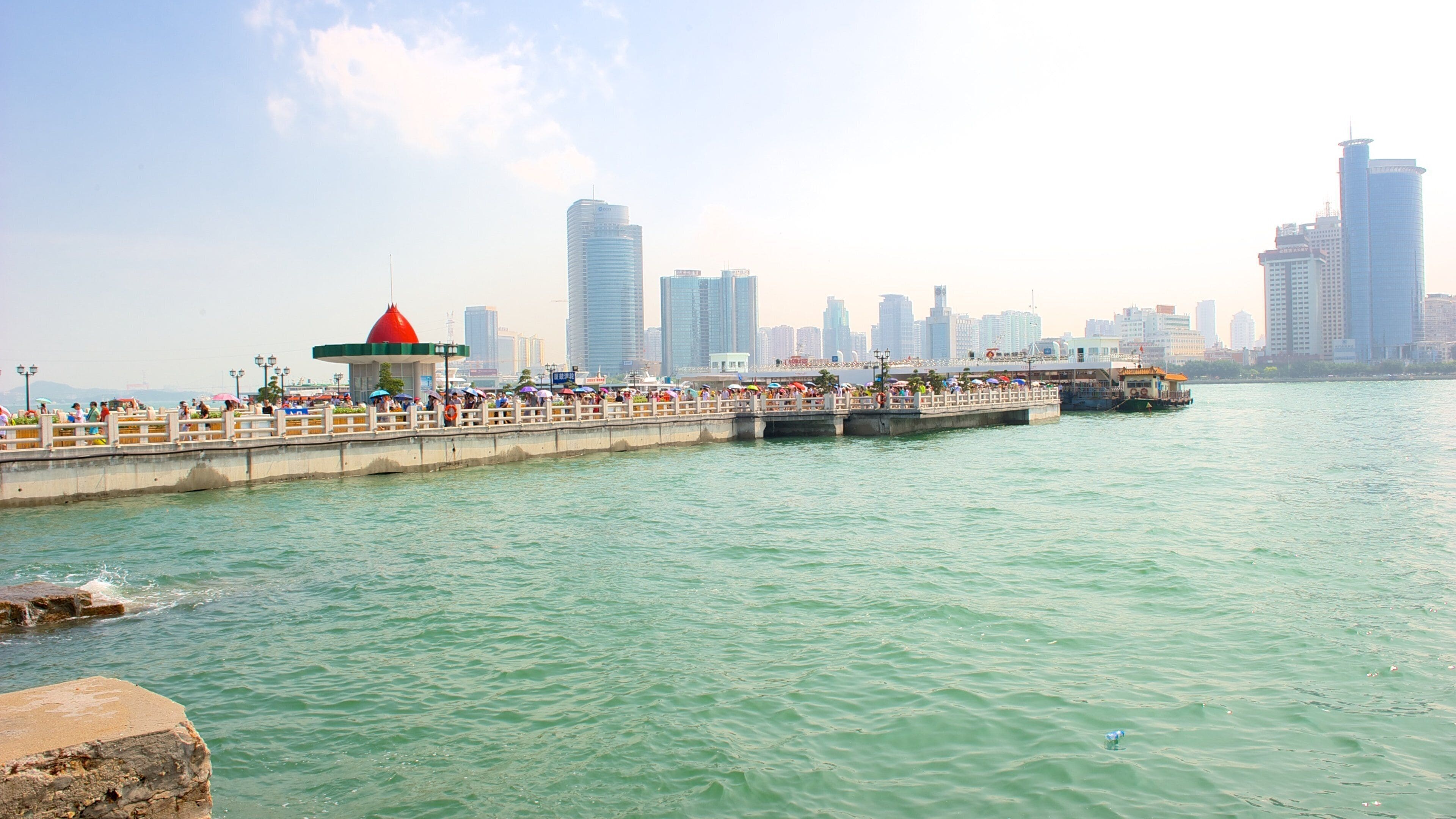 A waterfront promenade with a red-roofed pavilion, a pier extending into the water, and a city skyline in the background.