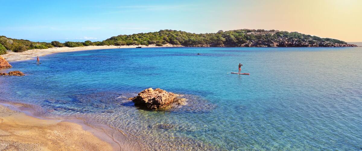 A clear coastal area with a sandy beach, calm blue waters, and a rocky outcrop in the foreground.