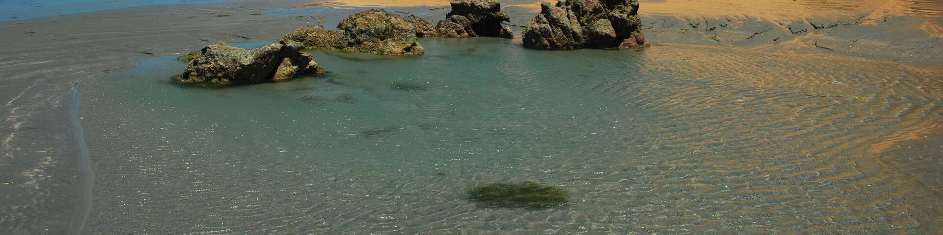 A beach with clear water, sand dunes, and rocky outcrops.