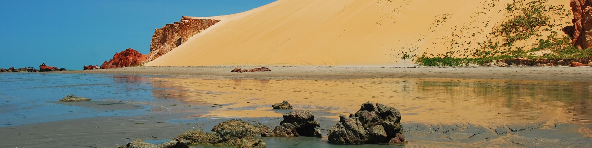Una playa con agua cristalina, dunas de arena y afloramientos rocosos.