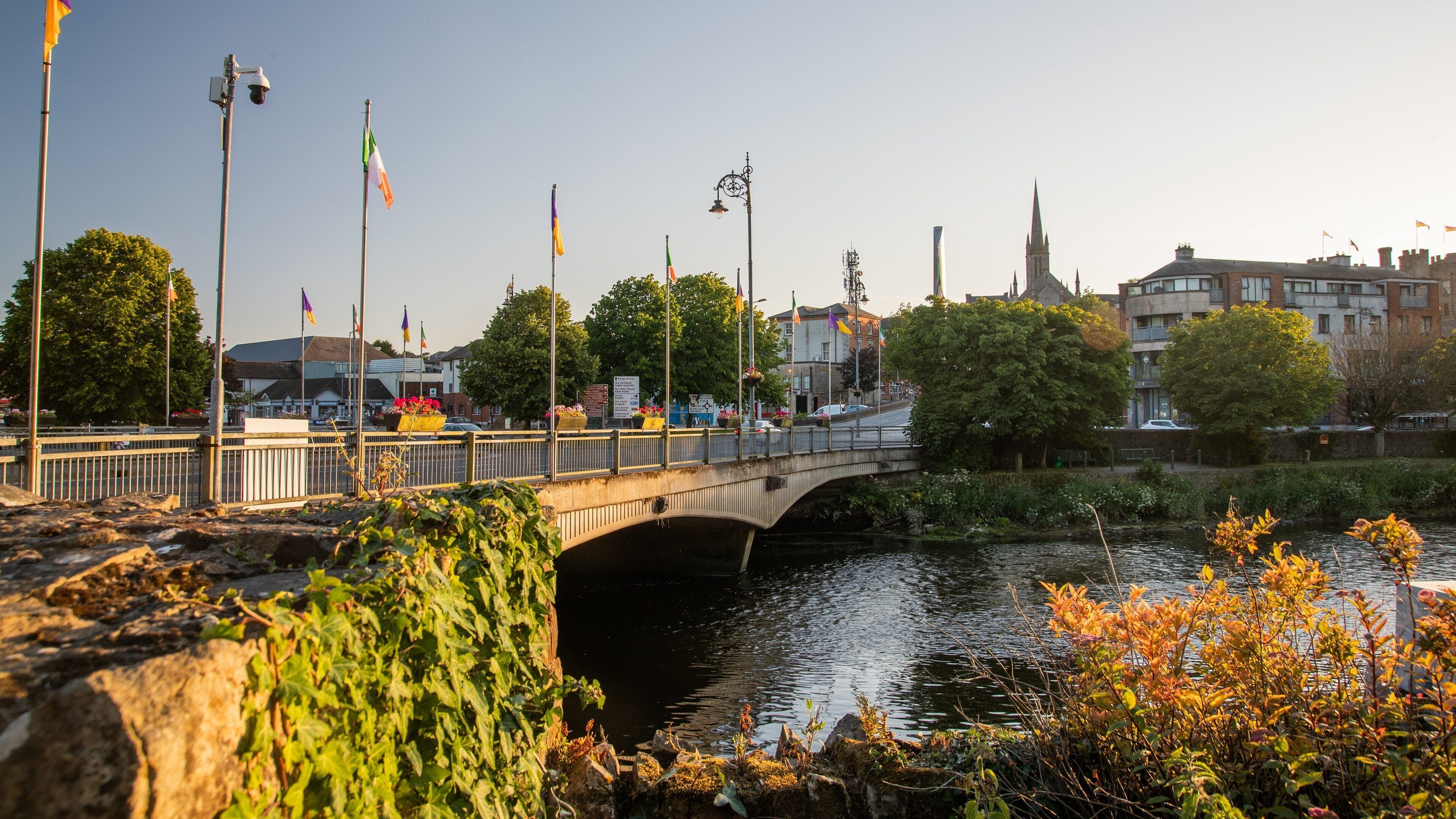 A stone bridge over a river with flags, trees, and buildings in the background.