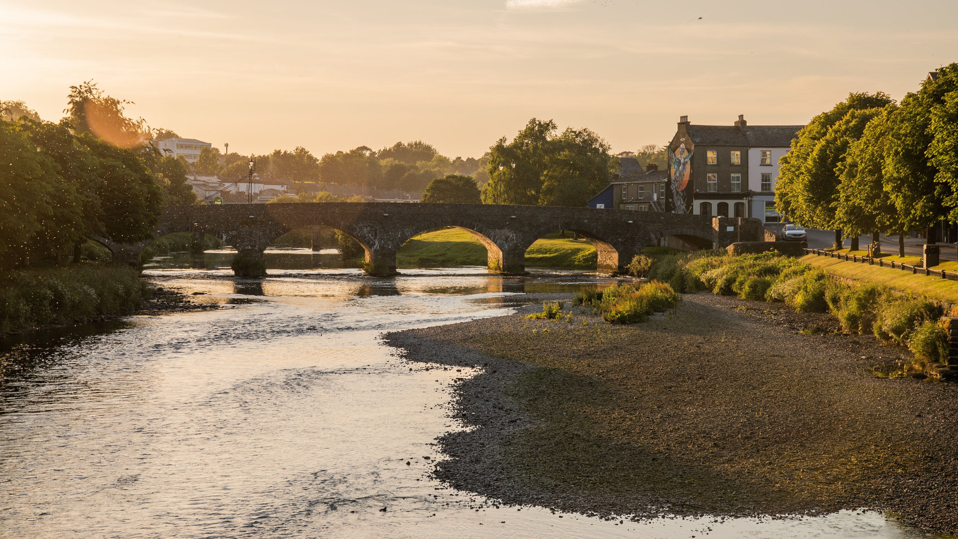 A stone bridge over a river with a grassy bank and buildings in the background.