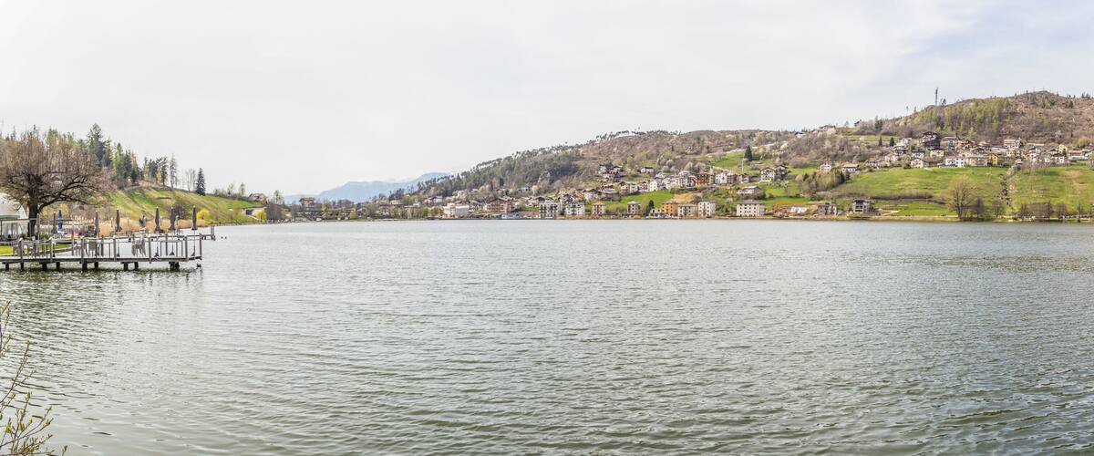 A calm lake with a dock on the left, surrounded by a hillside town with houses and greenery.