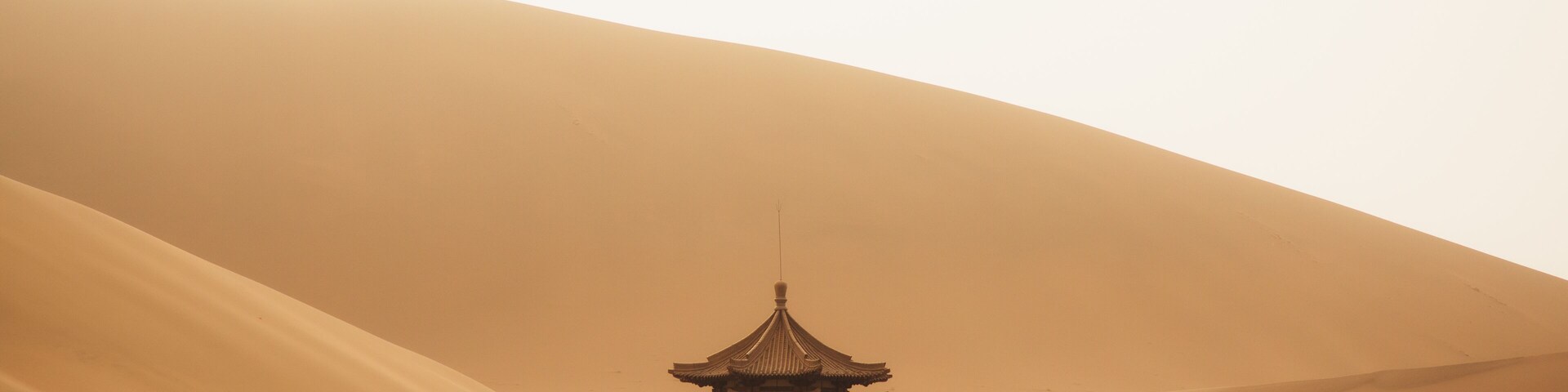 Una pagoda china tradicional con techos escalonados, rodeada de vegetación escasa y una gran duna de arena al fondo.