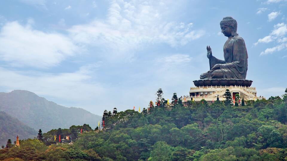 A large Buddha statue on a hill with a temple in the background and mountains in the distance.