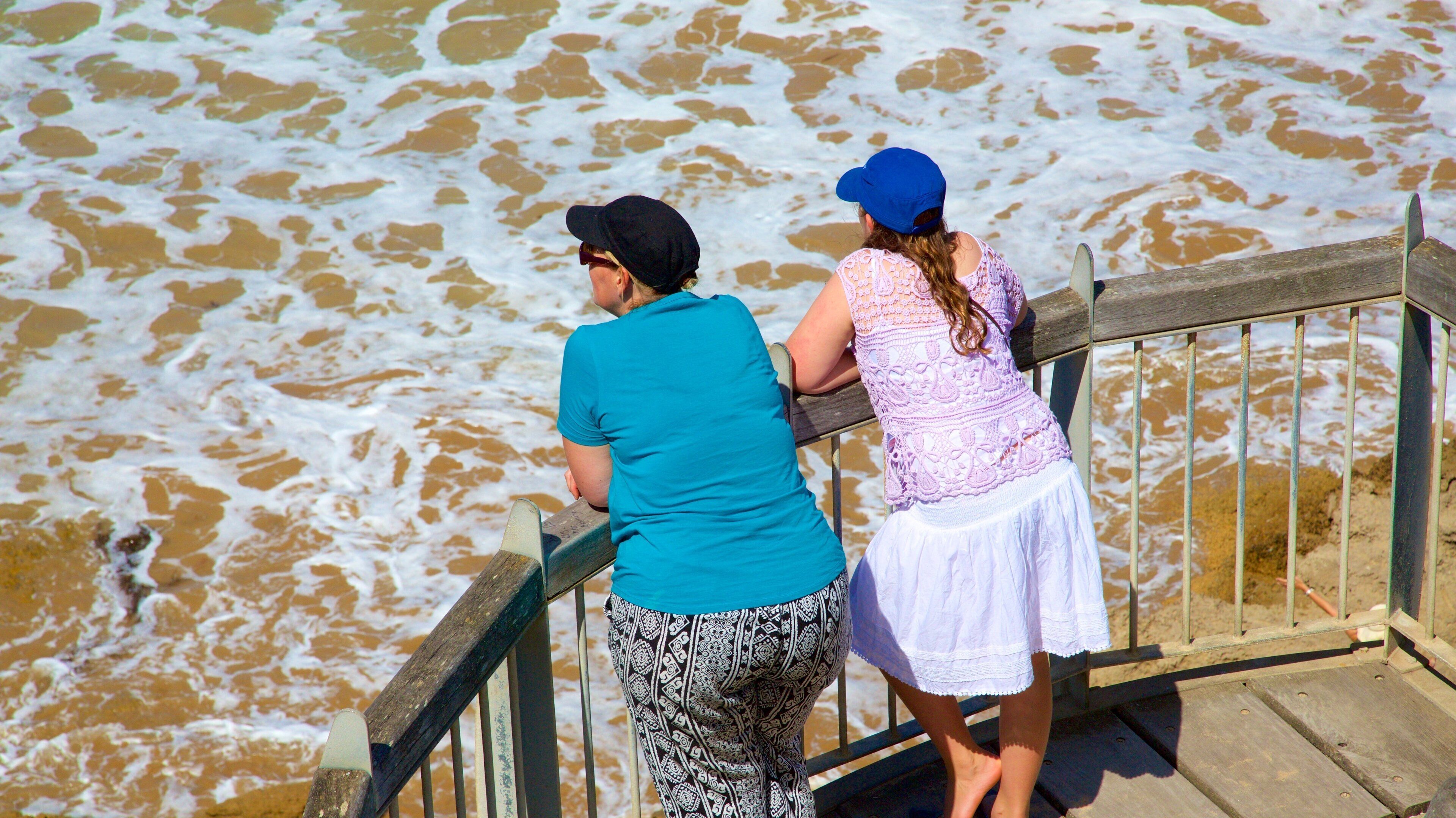 Two people on a wooden boardwalk overlooking a body of water with brownish foam.