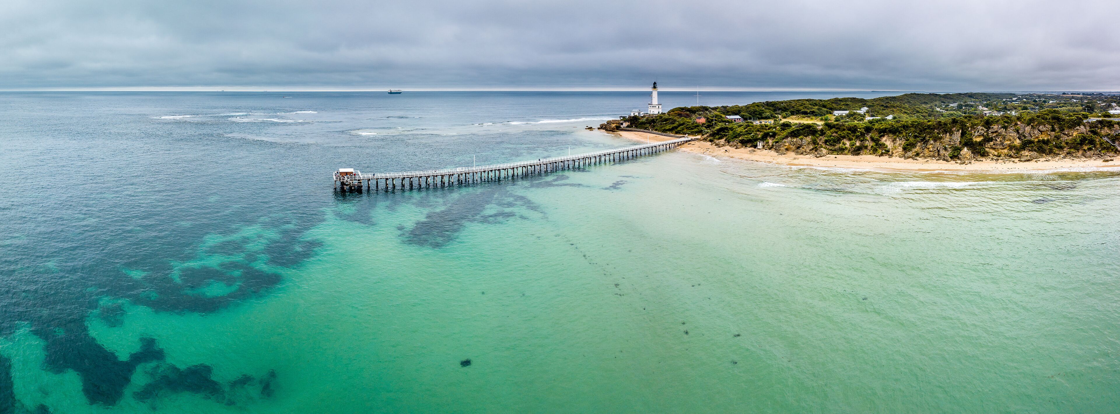 A coastal landscape with a lighthouse, a pier extending into the sea, and a beach with vegetation.