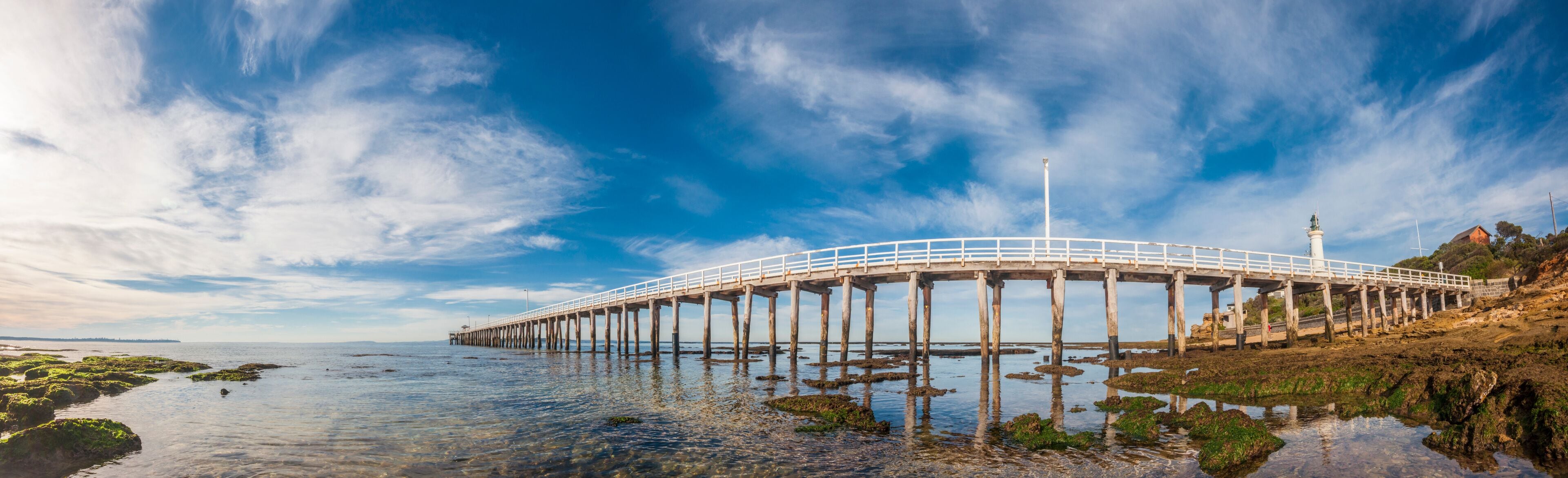 A coastal pier with a white railing, extending into calm waters with visible rocks and greenery.