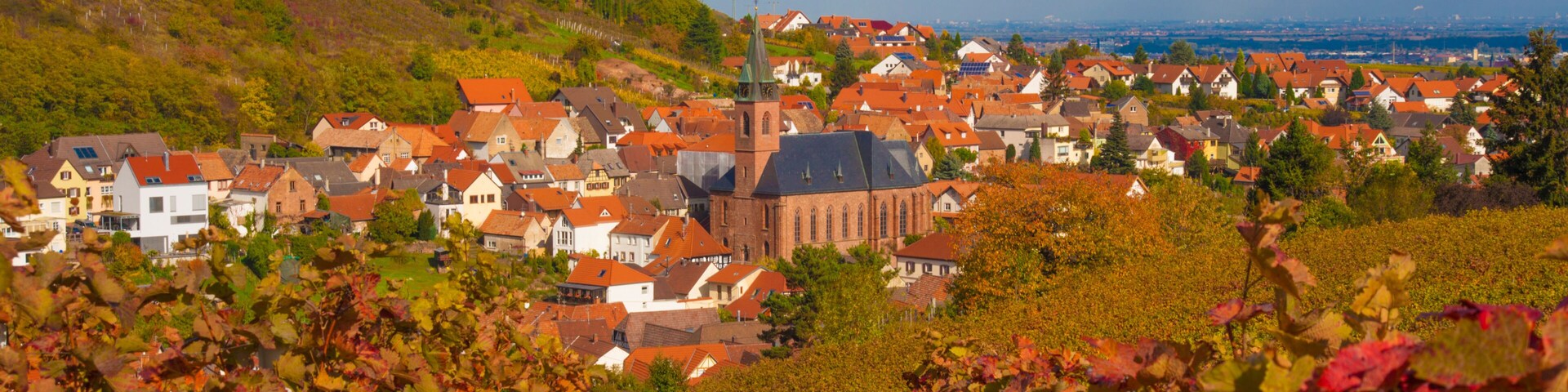 A town with red-roofed houses, a prominent church with a tall steeple, and a hillside in the background.