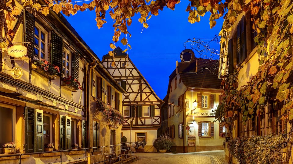 A cobblestone street lined with traditional European buildings, a sign for 'Friseur,' and a 'WEINSTUBE' establishment.