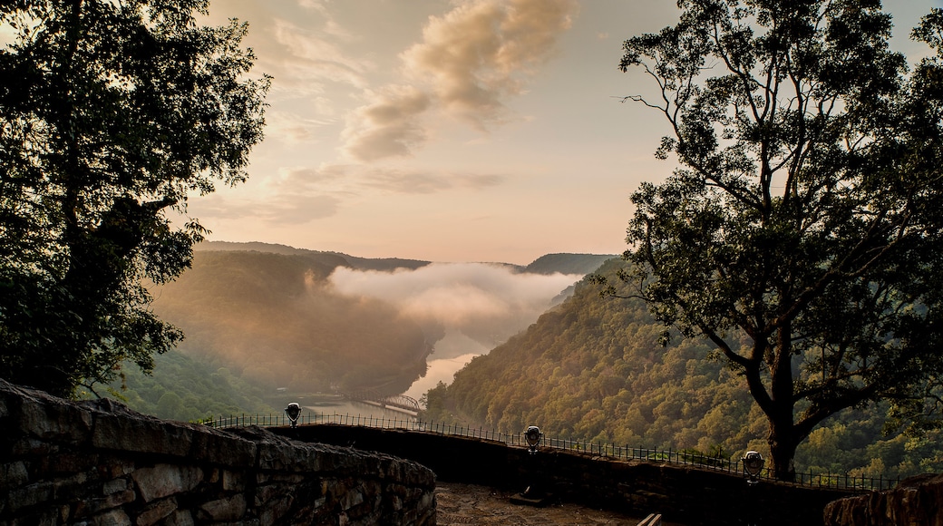Un mirador pintoresco con un muro de piedra, un banco de madera y vistas a un río con un puente y colinas cubiertas de niebla.